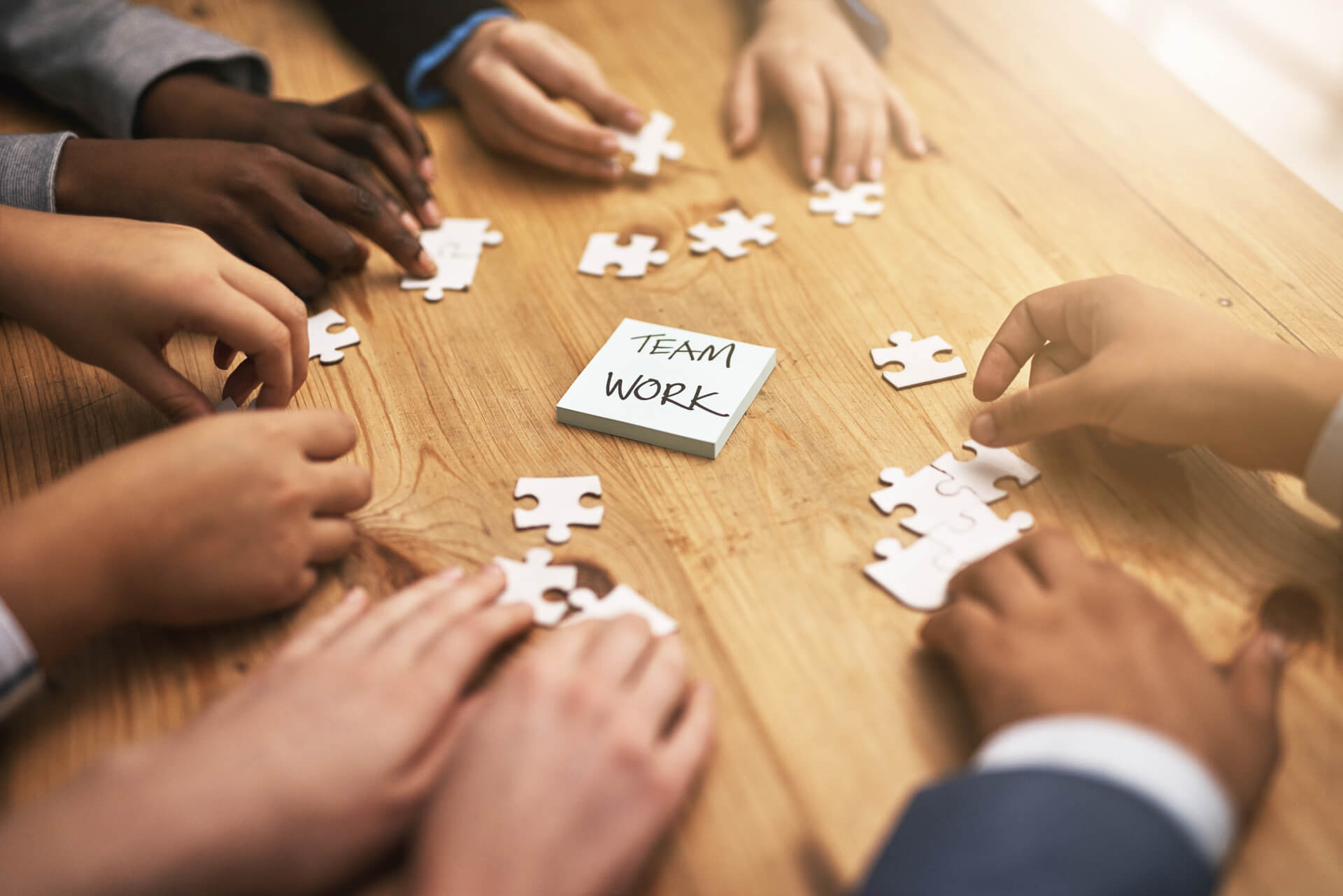 A close-up view of several hands collaborating over a wooden table, working on a jigsaw puzzle. In the center, a sticky note with the words "TEAM WORK" is prominently displayed among scattered puzzle pieces. The hands represent diverse skin tones, emphasizing teamwork and collaboration.