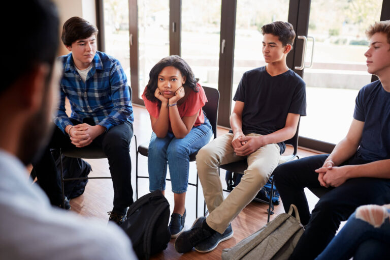 A group of four teenagers sitting in a circle in a bright room with large windows. One girl is sitting cross-legged, resting her chin on her hands with a concerned expression. The other three boys are sitting in their chairs, looking somewhat distracted. The boy in a plaid shirt and black pants is facing slightly towards the girl, while the others sit relaxed with their hands in their laps. Backpacks are resting on the floor beside them.